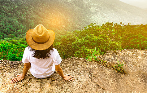 年轻的旅行妇女戴着帽子 坐在山崖顶上 心情放松 在度假时欣赏树林 蓝天和云彩的美丽景色 亚洲女人独自旅行天空孤独背包岩石游客森林图片