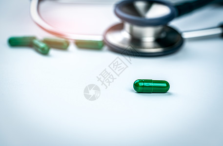 在模糊的听诊器和绿色胶囊药物背景下选择性地关注绿色胶囊药丸 医生白桌上的药物和听诊器 医疗保健和健康检查的概念 心脏病学图片