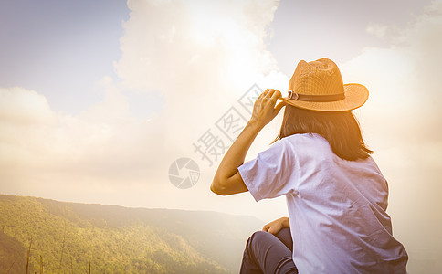 年轻的旅行妇女戴着帽子 坐在山崖顶上 心情放松 在度假时欣赏树林 蓝天和云彩的美丽景色 亚洲女人独自旅行女性孤独游客悬崖闲暇热带图片