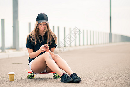 坐在滑板上时使用智能手机的年轻美丽的笑脸金发女孩金发女郎牛仔裤青少年运动木板女性电话滑冰短裤溜冰者图片
