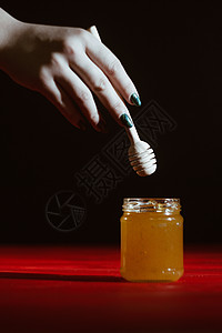 玻璃罐中的蜂蜜紧靠在红色木桌上的黑色背景上 用棒状蜂蜜勺倒蜂蜜花粉集装箱金子糖浆液体甜点玻璃作品食物橙子图片