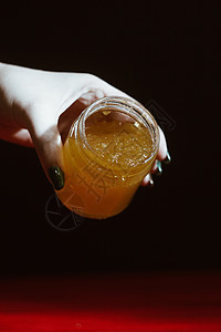 玻璃罐中的蜂蜜紧靠在红色木桌上的黑色背景上 用棒状蜂蜜勺倒蜂蜜生物花粉集装箱食物桌子作品养蜂业橙子蜂蜡杏仁图片