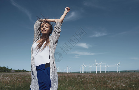 在风力涡轮机旁边 长发缠头发的女人风车资源燃料双臂力量可持续生活方式女士开襟衫自然界图片