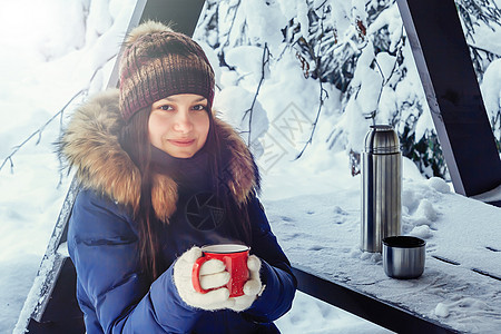 年轻女孩手里拿着一杯咖啡喝着热咖啡 在冬天雪覆盖的森林里的长椅上图片