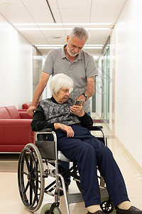 中年男子展示和帮助坐轮椅的95岁老人妇女如何使用现代移动电话 在轮椅上露面图片
