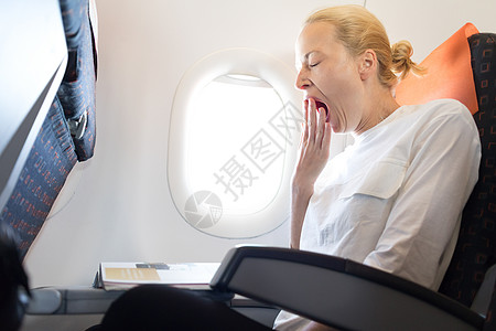 美丽的休闲白种女人在飞行杂志上阅读时在飞机上打呵欠图片