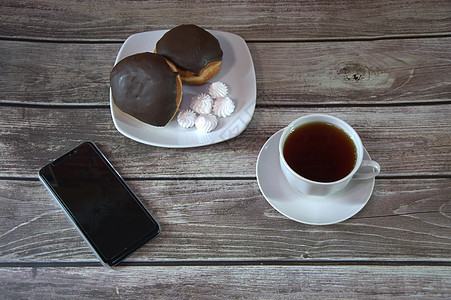 一杯红茶放在碟子上 盘子里放着两个涂有巧克力糖衣的甜甜圈和大块面包 放在智能手机旁边的木桌墙上 咖啡时间 休闲时光 特写厨房桌子背景图片