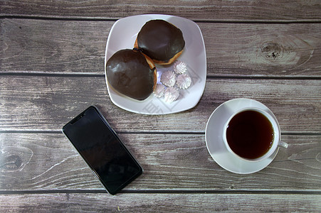 一杯红茶放在碟子上 盘子里放着两个涂有巧克力糖衣的甜甜圈和大块面包 放在智能手机旁边的木桌墙上 咖啡时间 休闲时光 特写杯子厨房图片