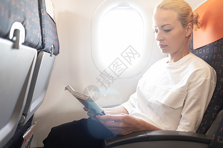 在飞机上阅读飞行杂志的女人 女性旅客坐在客舱阅读 阳光透过飞机窗旅行女郎窗户航班乘客木板空气金发女孩经济图片