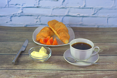 一杯黑咖啡放在带勺子的碟子上 一盘羊角面包和柠檬片加黄油放在木桌上 特写美食甜点小吃食物营养咖啡活力早餐桌子杯子图片