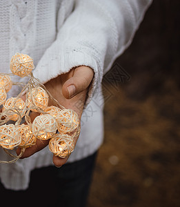 手掌上的光 女手拿着一个带柳条圆球的花环 身穿白色毛衣 手里拿着圣诞灯的女孩 温暖的新年气氛 柔和的焦点手指细绳黑板杯状面包风格图片