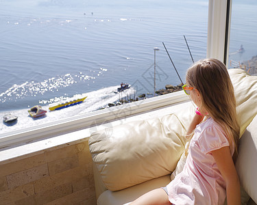 一个小女孩坐在窗边看着大海 从窗户到大海的景色 从度假村的阳台看海景的年轻女子的后视图图片