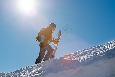 滑雪运动员在太阳尼日与雪板一起爬上山中山 滑雪和冬季运动风镜行动娱乐男人蓝色粉末滑雪板季节单板假期图片