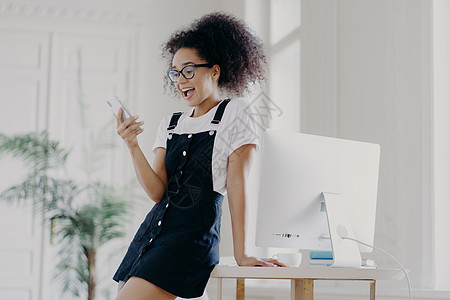 室内拍摄的年轻 没有经验的非裔美国女性戴着眼镜 白色 T 恤和 sarafan 使用手机在线聊天 在电脑桌旁摆姿势图片
