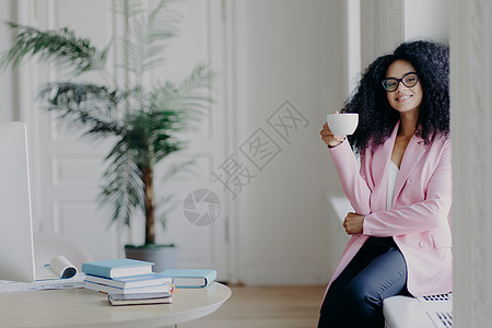 积极的非洲裔美国女商务人士坐在窗台前 拿着一杯咖啡 穿着正式的衣服 在桌面附近摆姿势 摆着书本 背景是绿色室内植物 休息了图片