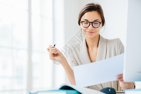 专注的成功女商务人士在纸上专心地看着 研究合同条款 拿着笔 写在文件中 穿着正式 在桌面上摆姿势 反对白色宽敞的内部图片