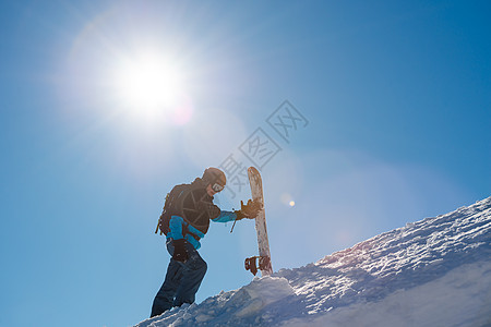 滑雪运动员在太阳尼日与红雪板一起爬上山中山顶 滑雪和冬季运动下坡男人娱乐行动风镜季节背包单板粉末旅行图片