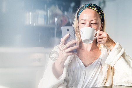 家里美丽的女性 穿着白浴袍感到舒服 自己花点时间 喝早咖啡及早上在手机设备上读新闻   info饮料房子技术女孩杯子厨房工作成人图片