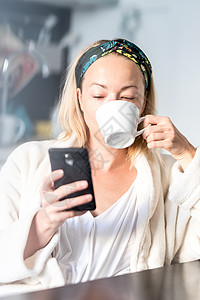 家里美丽的女性 穿着白浴袍感到舒服 自己花点时间 喝早咖啡及早上在手机设备上读新闻   info厨房电话乐趣女孩浴衣技术互联网阅图片