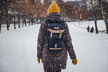 快乐的年轻欧洲女人戴着黄色针织帽 戴着手套 背着背包 在大雪中笑着离开 从后面看 复古效果 模糊降雪黑发幸福天气薄片青年外套女士图片