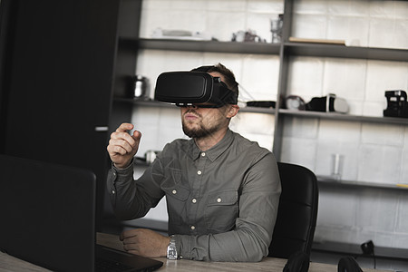 留着胡须的年轻男子戴着虚拟现实眼镜坐在电脑前 试图触摸或轻敲他所看到的东西 现代技术 未来技术的概念娱乐活动风镜工作室展示工具男图片