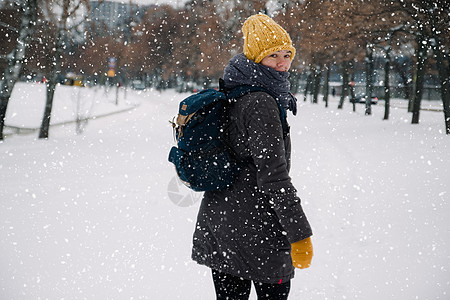 快乐的年轻欧洲女人戴着黄色针织帽 戴着手套 背着背包 在大雪中笑着离开 从后面看 复古效果 模糊毛衣薄片眼睛帽子青年天气微笑自由图片