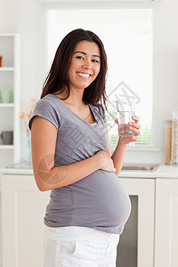 怀孕的孕妇在站立时拿着一杯水喝着女孩父母享受头发玻璃灰色女士家庭厨房亮度图片