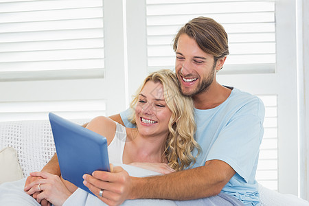 使用平板电脑在毯子下坐在沙发上坐着的微笑临时夫妇女性拥抱滚动男性空闲触摸屏房子客厅长椅亲密感图片