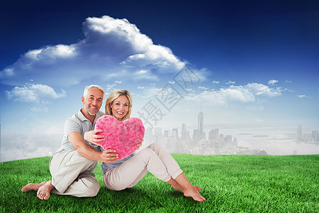 幸福的情侣坐着 抱着心脏枕头的综合形象软垫亲密感休闲男人大厦金发头发微笑金发女郎拥抱图片