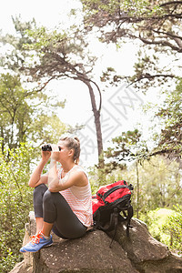 坐在岩石上看望望远镜的金发登山者荒野越野女士闲暇石头风景林地农村远足冒险图片