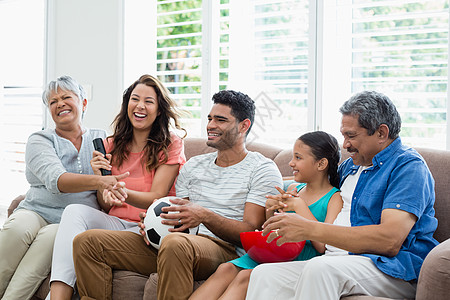 快乐的多代家庭在客厅的电视上观看足球比赛运动混血孩子祖母女士爆米花零食父亲女儿房子背景图片