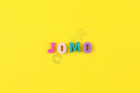 在柔和的黄色背景上用五颜六色的木制字母缩写词 JOMO JOMO  错过的快乐 反对 选择 社会问题 数字排毒 平躺 复制空间 图片