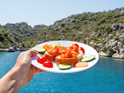 地中海美食-面包上新鲜煮熟的虾和蔬菜 在美丽的大海泻湖背景上拿着盘子的手 海鲜配黄瓜和西红柿图片