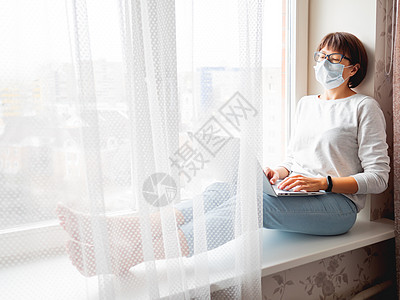 身戴医疗面具的妇女在家工作 坐在窗户上口罩窗台远程教育键盘成人封锁教育按钮技术自由职业者图片