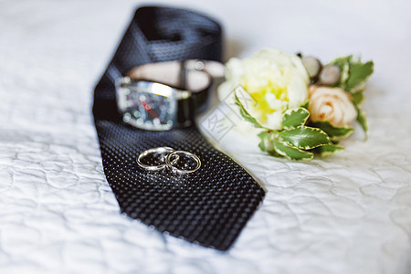 一对金色结婚戒指 黑色领带上镶有钻石 背景上有一朵玫瑰花和一块手表的胸花 婚礼上新郎的传统配饰图片