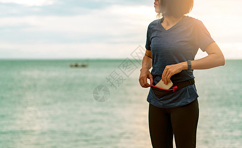 年轻亚洲女性在跑步前用腰包装智能手机海岸腕带旅行乐队闲暇有氧运动海滩天空假期手腕图片