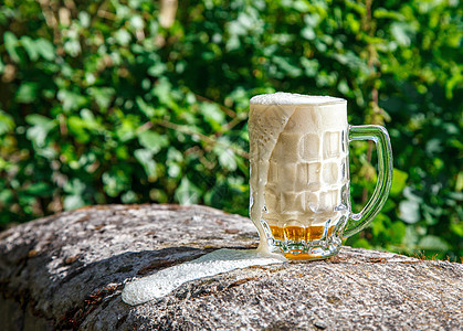 杯子上装着啤酒的玻璃杯 站在大石头上绿色阳光气泡公园玻璃森林树木黄色泡沫日光图片
