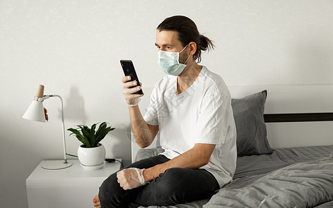 由于冠状病毒的流行 戴医用口罩的男子正在家里坐在床上打电话 大流行期间的远程工作 在 COVID19 检疫概念期间待在家里学家成图片