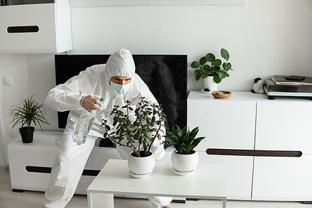 身着防护服 医疗面具和橡皮手套的人在家中起居室照看用喷雾器喷洒的植物 同时对COVID19进行冠状病毒检疫面具防护口罩男人安全技图片