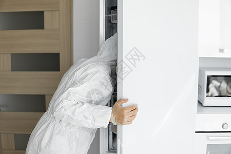 在冠状病毒期间 身穿白色防护服 戴着医用口罩的男子盯着家里白色厨房的冰箱 呆在家里 享受在家做饭的乐趣 COVID19 全球大流图片