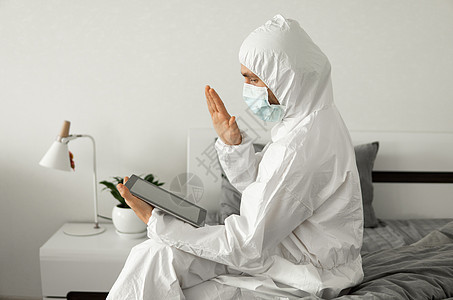 身穿保护性白色西装和医疗面具的人在家用笔记本电脑在床上工作 并因冠状病毒流行而向朋友或商业伙伴打招呼成人防护服面具药品互联网技术图片