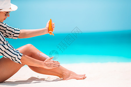 年轻女子在白沙滩上用防晒霜申请假期奶油身体日光浴闲暇蓝色防晒晴天海滩系数图片