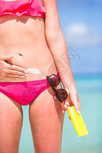 年轻女子在白沙滩上用防晒霜申请身体女性女士旅行日光浴闲暇瓶子防晒奶油蓝色图片