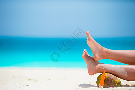 白沙滩上女性棕色光滑的双腿 加上太阳淇淋和椰子热带假期紫外线晒黑海滩棕褐色防晒霜防晒异国休息图片