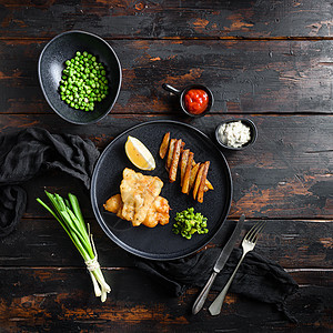 由油炸鱼 薯条 木薯豆和塔塔酱组成的传统英国快餐热菜 在酒吧顶视角广场的旧黑木桌上用黑色盘子供应文字材料空间图片