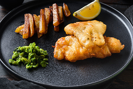 由油炸鱼 薯条 木薯豆和塔塔酱组成的传统英国式热菜 在酒吧边的旧黑木板上用黑色盘子喂食图片