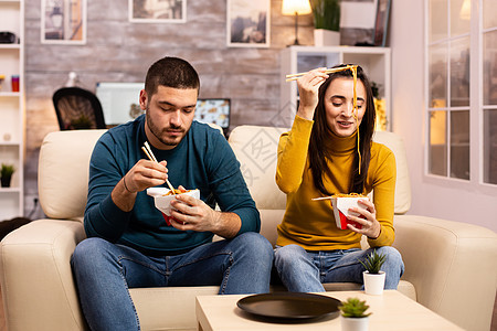 在现代舒适的客厅里 夫妻在看电视时 正在享受外送面条成人食物女士女朋友房子男人筷子沙发手表长椅图片