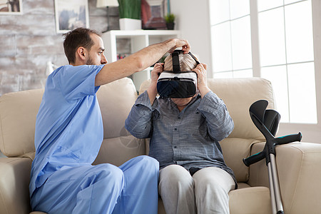 年轻医生给年长妇女戴上虚拟现实眼镜的青年医生病人疗养院照顾者头发护士沙发拐杖老年女士疾病图片