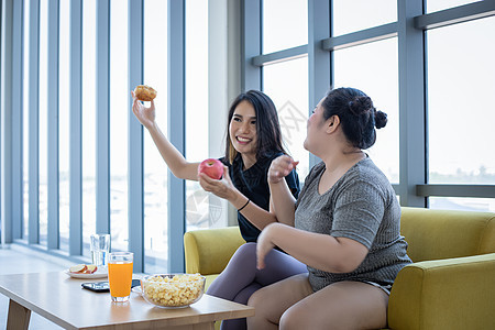超重妇女和亚洲女孩享受在家沙发上吃食物肥胖手表成人垃圾女士营养长椅小吃女性电视图片
