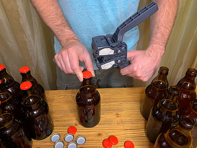 在家酿造的手工艺啤酒 男人特写棕色玻璃啤酒瓶 用红色冠盖在木制桌上的塑料罩子液体软木工具密封草稿盖帽机器酿酒师补给品金属图片